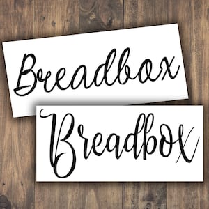 May include: Two white rectangular labels with the word "Breadbox" in elegant black cursive script. The labels are placed on a wooden background, creating a contrast between the white and the dark wood.