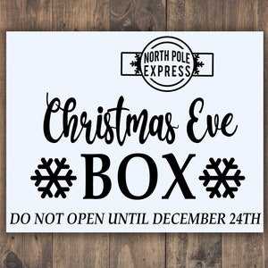 May include: A white sign with black text that reads "Christmas Eve BOX" with snowflakes on either side of the word "BOX". The sign also has a black and white stamp that reads "NORTH POLE EXPRESS" and the text "DO NOT OPEN UNTIL DECEMBER 24TH".