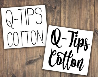 Puede incluir: Dos letreros rectangulares blancos con las palabras "Q-TIPS COTTON" en letras negras. Un letrero tiene una fuente simple, mientras que el otro tiene una escritura más estilizada. Los letreros están sobre un fondo de madera.