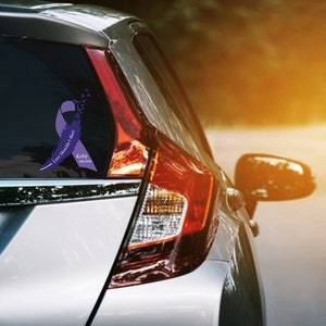 Puede incluir: Una cinta morada con plumas y el texto "Love Should've Last Forever" y "Kelly 1965-2018" se muestra en la ventana trasera de un coche plateado.