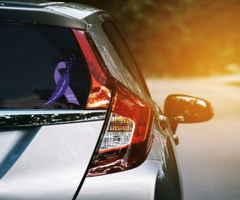Puede incluir: Una cinta morada con plumas y el texto "Love Should've Last Forever" y "Kelly 1965-2018" se muestra en la ventana trasera de un coche plateado.