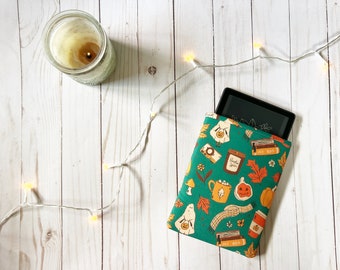 Puede incluir: Una funda para lector electrónico de color verde azulado con un estampado otoñal de calabazas, fantasmas, libros y tazas. Una vela encendida en un tarro de cristal y luces de hadas sobre una superficie de madera blanca.