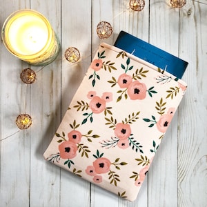 Könnte beinhalten: Eine rosa-weiße Blumen-Buchhülle mit einem blauen Buch darin. Die Buchhülle liegt auf einer weißen Holzoberfläche mit einer Lichterkette.