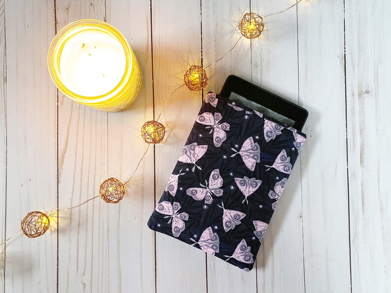 K&ouml;nnte beinhalten: Eine schwarze E-Reader-H&uuml;lle mit einem pinkfarbenen und wei&szlig;en Schmetterlingsmuster. Die H&uuml;lle liegt auf einer wei&szlig;en Holzoberfl&auml;che mit einer Lichterkette.
