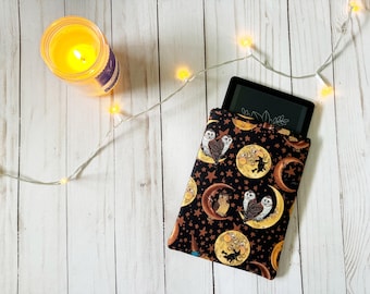 May include: A black e-reader sleeve with a moon and owl pattern. The sleeve is made of fabric and has a zipper closure. The sleeve is on a white wooden surface with string lights.