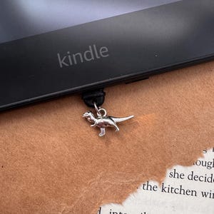 Puede incluir: Un dije de dinosaurio plateado cuelga de un lector electrónico Kindle negro. La palabra "kindle" está impresa en el dispositivo. El fondo es una superficie marrón con texto de libro.