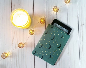 May include: A green e-reader case with a moon and star pattern. The case is on a white wooden surface with a string of lights.