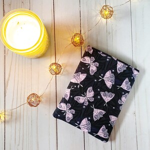 K&ouml;nnte beinhalten: Ein schwarzer Stoff-Notizbuchumschlag mit einem rosa-wei&szlig;en Schmetterlingsmuster. Der Umschlag liegt auf einer wei&szlig;en Holzoberfl&auml;che mit einer Lichterkette.