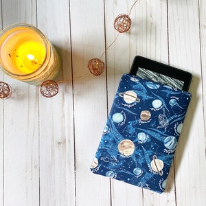 May include: A blue and black e-reader cover with a galaxy print featuring planets and stars. The cover is made of fabric and has a zipper closure.