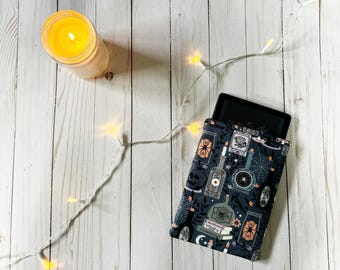May include: A black and gray e-reader sleeve with a spooky, vintage-inspired print featuring potion bottles, candles, and other witchy elements. The sleeve is on a white wooden surface with a string of fairy lights.