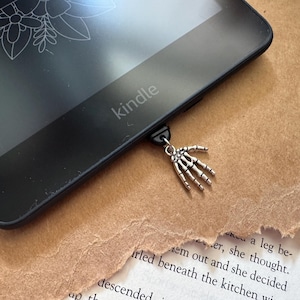 Könnte beinhalten: Ein schwarzer Kindle E-Reader mit einem silbernen Skelett-Hand-Lesezeichen, das am unteren Rand befestigt ist. Das Wort "Kindle" ist auf dem E-Reader sichtbar.