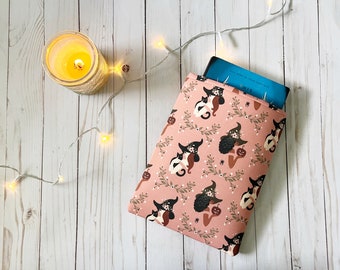 Könnte beinhalten: Eine rosa Buchhülle mit einem Hexen- und Katzenmuster. Die Hülle ist mit einem blauen Buch gefüllt, auf dessen Umschlag der Text "This One" steht.