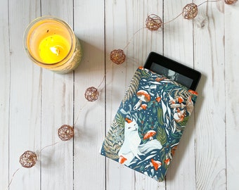 Puede incluir: Una funda para lector electrónico de color azul y verde con un patrón de zorro blanco y setas rojas. La funda está sobre una superficie de madera blanca con una cadena de luces de cobre.