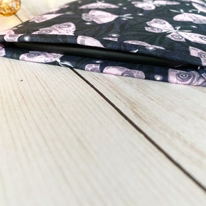 K&ouml;nnte beinhalten: Eine schwarze und rosafarbene Tableth&uuml;lle aus Stoff mit einem Muster aus Motten. Die H&uuml;lle ist zusammengefaltet und das Tablet ist nicht sichtbar.