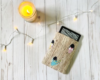 Puede incluir: Una funda de lector electrónico marrón con un patrón de veta de madera y búhos coloridos asomándose por los agujeros. La funda está sobre una superficie de madera blanca con una cadena de luces blancas.