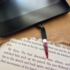 May include: A black Kindle e-reader with a bookmark attached. The bookmark is a small, red and black knife with a red handle.