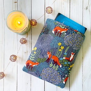 Könnte beinhalten: Eine blaue Buchhülle mit einem Fuchs- und Blumenmuster. Die Hülle ist aus Stoff und hat ein Buch darin. Die Hülle liegt auf einer weißen Holzoberfläche mit einer Lichterkette.