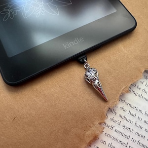 Peut inclure: Un marque-page en forme de crâne d'oiseau argenté avec des détails complexes pend d'une liseuse Kindle noire. Le marque-page est en forme de crâne d'oiseau avec un long bec.