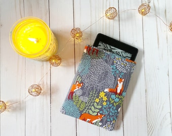 May include: A gray and blue fabric e-reader cover with a woodland pattern featuring orange foxes, green trees, and white flowers. The cover is lying on a white wooden surface with a string of lights.