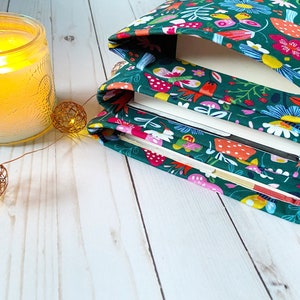 May include: A stack of books covered in green fabric with a colorful floral and mushroom pattern. The fabric has a white border around the edges. The books are sitting on a white wooden surface.