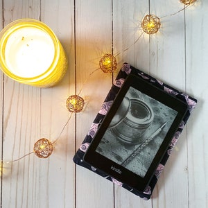 K&ouml;nnte beinhalten: Ein schwarzer Kindle E-Reader mit einer gemusterten H&uuml;lle mit rosa und schwarzen Augen. Der E-Reader liegt auf einer wei&szlig;en Holzoberfl&auml;che mit einer Lichterkette.