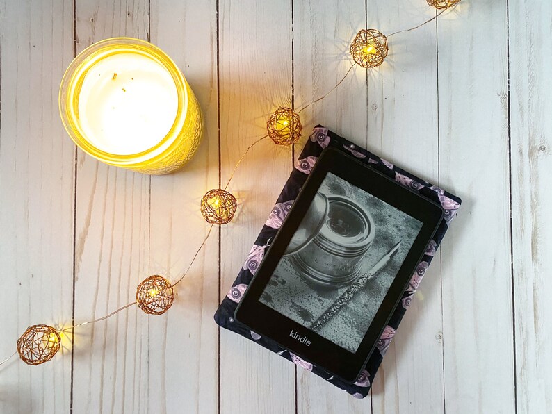 K&ouml;nnte beinhalten: Ein schwarzer Kindle E-Reader mit einer gemusterten H&uuml;lle mit rosa und schwarzen Augen. Der E-Reader liegt auf einer wei&szlig;en Holzoberfl&auml;che mit einer Lichterkette.