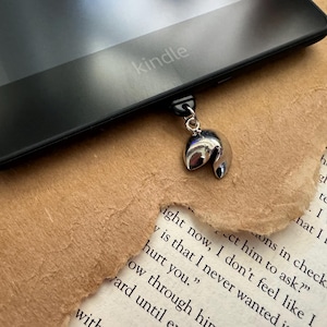 Puede incluir: Un amuleto de galleta de la fortuna plateado sujeto a un lector electrónico Kindle negro. El amuleto cuelga de la parte superior del lector electrónico.