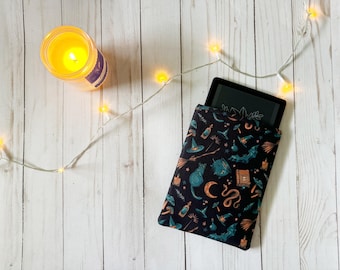 May include: A black e-reader in a black and green fabric case with a witchy pattern. The pattern includes a black cat, a cauldron, a snake, and a witch's hat. The case is lying on a white wooden surface with a string of white lights.