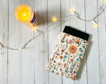 May include: An e-reader in a floral print case with orange, pink, and blue flowers. A lit candle in a glass jar and a string of lights are also in the image. The background is a white wooden surface.