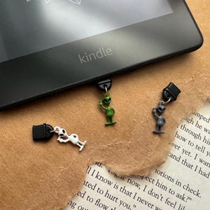 May include: Three alien-themed bookmark clips. One is green, one is grey, and one is white. The clips are attached to a black Kindle e-reader.
