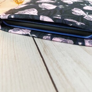 K&ouml;nnte beinhalten: Eine schwarze Stoffh&uuml;lle mit einem rosa-wei&szlig;en Schmetterlingsmuster, die f&uuml;r einen E-Reader konzipiert ist. Die H&uuml;lle liegt auf einer hellen Holzoberfl&auml;che.