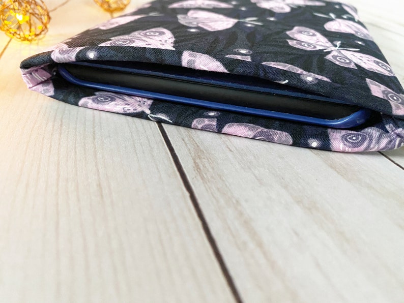 K&ouml;nnte beinhalten: Eine schwarze Stoffh&uuml;lle mit einem rosa-wei&szlig;en Schmetterlingsmuster, die f&uuml;r einen E-Reader konzipiert ist. Die H&uuml;lle liegt auf einer hellen Holzoberfl&auml;che.