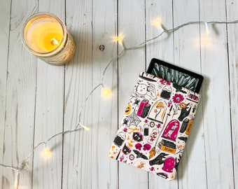 May include: A white e-reader sleeve with a pink, black, and orange Halloween-themed print. The print features various spooky and magical items, including skulls, bats, potions, and a crystal ball.