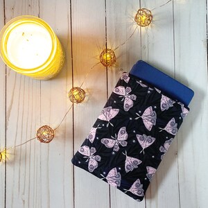 K&ouml;nnte beinhalten: Ein schwarzer und pinkfarbener Buchbezug aus Stoff mit Schmetterlingsmuster und einem blauen Buch darin. Der Bezug liegt auf einer wei&szlig;en Holzoberfl&auml;che mit einer Lichterkette.
