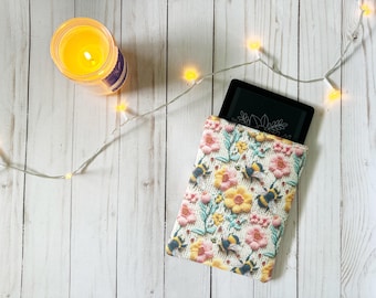 May include: A white e-reader case with a floral pattern featuring bees and flowers. The case is made of fabric and has a zipper closure.