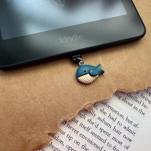 May include: A blue whale bookmark with a white belly and a black eye, attached to a black Kindle e-reader.