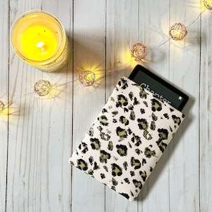 Puede incluir: Una funda para lector electrónico de color rosa y dorado con estampado de leopardo con un lector electrónico negro en su interior. La funda está sobre una superficie de madera blanca con una cadena de luces.