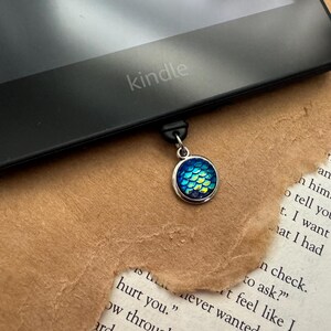 May include: A silver bookmark with a blue and green iridescent mermaid scale charm. The bookmark is attached to a Kindle e-reader.