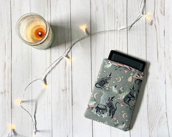 May include: A green and black e-reader cover with a witchy pattern featuring black cats, cauldrons, and stars. The cover is on a white wooden surface with a string of white lights.