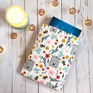 Könnte beinhalten: Eine weiße Buchhülle mit einem Blumenmuster mit Bienen, Bienenstöcken und Blumen. Die Hülle ist aus Stoff und hat ein blaues Buch im Inneren.