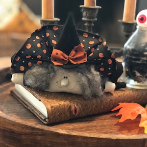 May include: A Halloween-themed decorative witch doll with a black hat and a bow, resting on a leather-bound book. The witch's hat is adorned with orange pumpkins. The scene includes candles and a decorative tray, evoking a festive autumn atmosphere.
