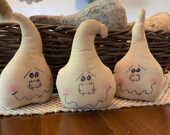 May include: Three beige fabric ghost-shaped decorations with stitched-on eyes and mouths. The ghosts are sitting on a woven placemat on a wooden table.