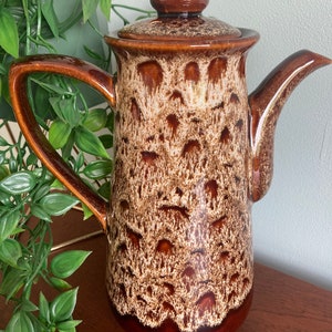 May include: A brown and white ceramic teapot with a mottled glaze. The teapot has a rounded lid and a spout.