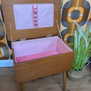 May include: A rectangular wooden sewing box with four tapered legs and a hinged lid. The interior is lined with quilted pink fabric, featuring a matching pink fabric panel on the underside of the lid with a vertical row of elasticised loops.