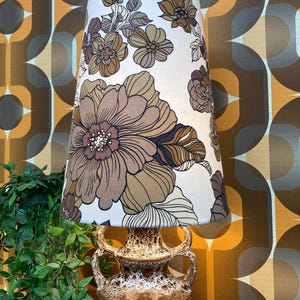 May include: A table lamp featuring a tall, tapered fabric lampshade with a brown and cream floral pattern. The base is made of ceramic with a speckled brown and white glaze, characterised by a wide body and two symmetrical arched handles.
