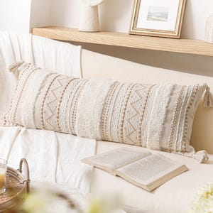 May include: A rectangular lumbar pillow featuring a cream and beige woven fabric with textured vertical stripes, geometric zigzag patterns, and small tufted details. The pillow is finished with decorative tassels at each of the four corners.