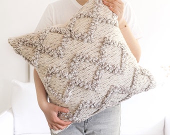 May include: A cream-colored throw pillow with a textured, woven design. The pillow has a geometric pattern with raised bumps and a subtle sheen.
