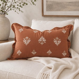 May include: A rectangular terracotta-colored throw pillow featuring a ruffled edge and a repeating pattern of embroidered white floral sprigs. The pillow is positioned on a light-colored sofa, showcasing a soft texture and detailed stitching.