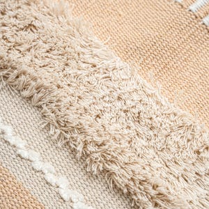 May include: Close-up of a woven textile with a textured, fringed design. The rug features a neutral color palette of beige and cream, with a pattern of stripes and fringed edges. The texture appears soft and inviting.