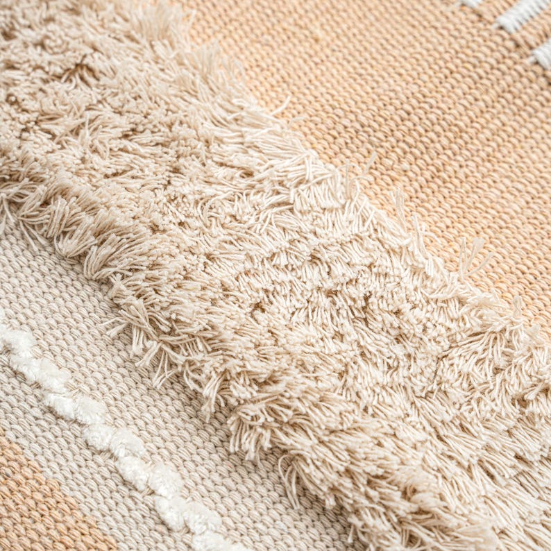 May include: Close-up of a woven textile with a textured, fringed design. The rug features a neutral color palette of beige and cream, with a pattern of stripes and fringed edges. The texture appears soft and inviting.